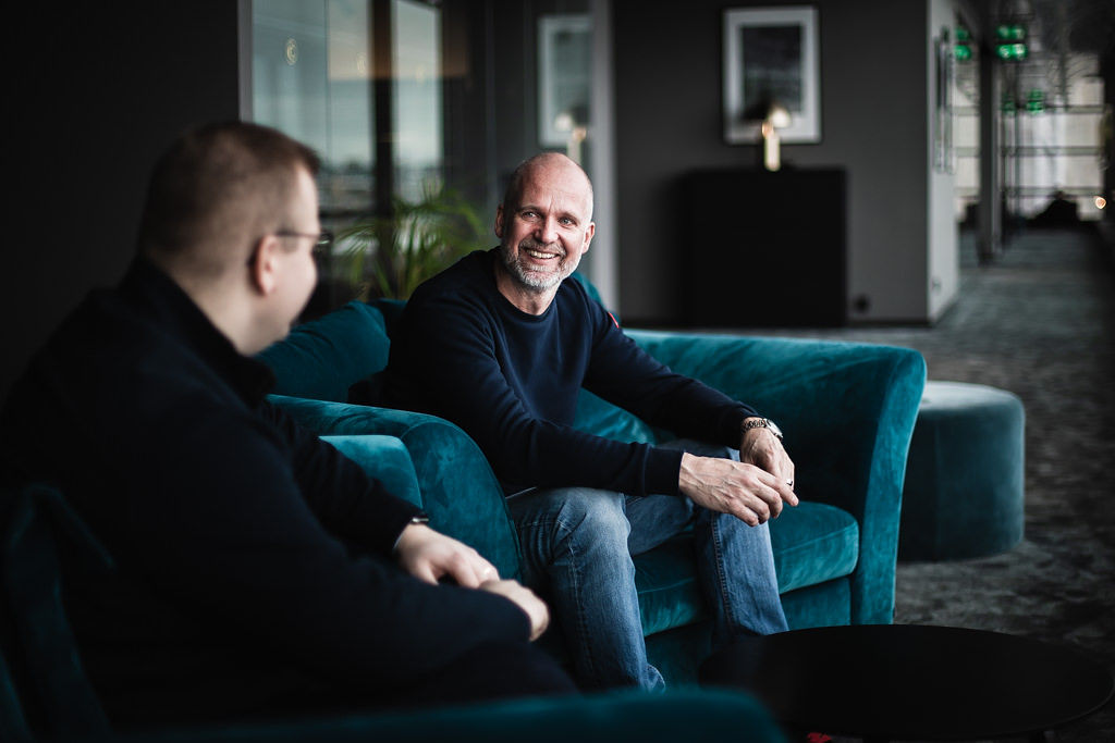 Joakim, VD på elastx, sitter i en mjuk fåtölj i en ljus och mysig korridor med ett leende medan han samtalar med en kollega.